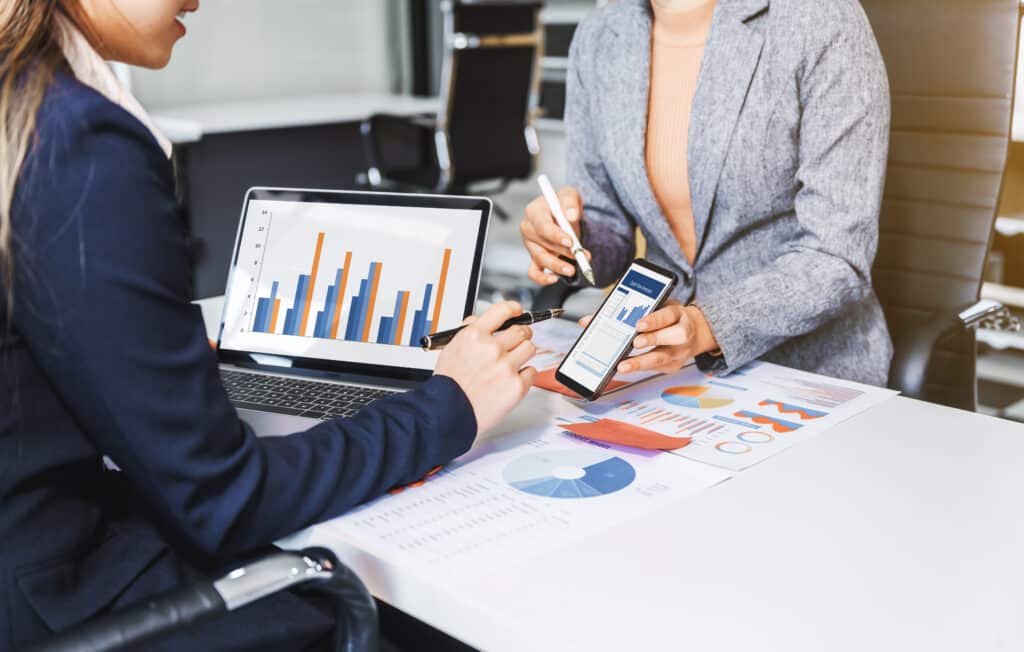 Two female business professionals in suits sit at desk, engaged in strategic discussion. They analyze financial planning, risk management, corporate growth while reviewing reports and market trends.